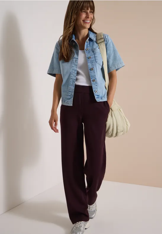 Femme souriante en pantalon bordeaux large, top blanc, veste courte en jean et baskets.