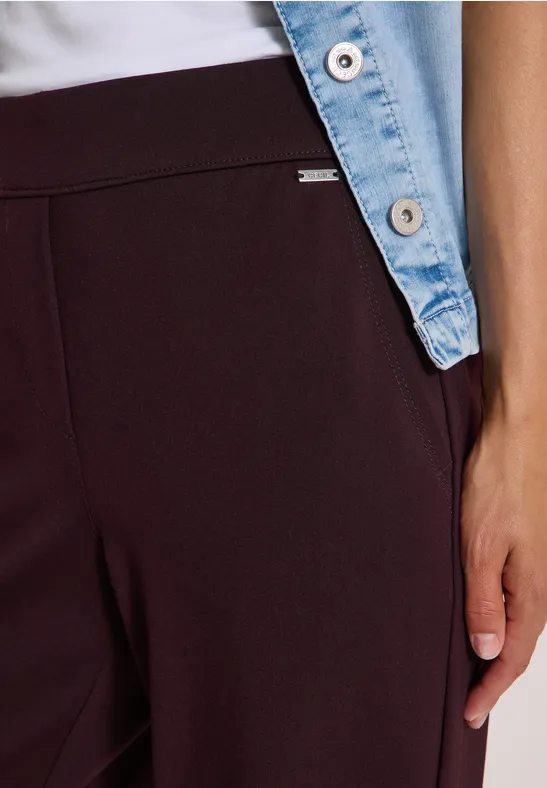 Gros plan sur une personne en pantalon bordeaux, haut blanc et veste en jean, main sur la cuisse