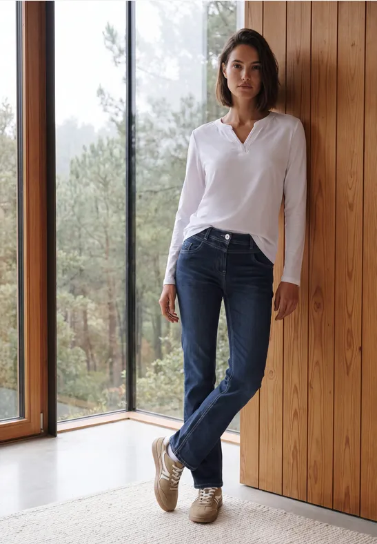 Eine Frau steht lässig in Jeans und weißer Bluse an einer Holzwand neben einem Fenster mit Waldblick.