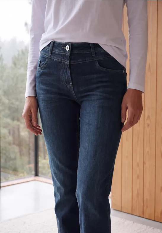 Eine Person in dunkelblauen Jeans und weißem Oberteil steht vor einem Fenster neben einer Holzverkleidung.