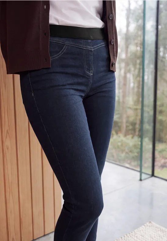 Eine Person trägt dunkle Jeans und steht vor einem Fenster neben einer Holzverkleidung.