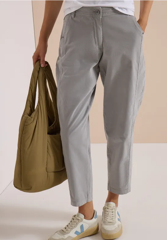 Person trägt graue, lockere Hose, weiße Sneakers und hält eine olivgrüne Umhängetasche.