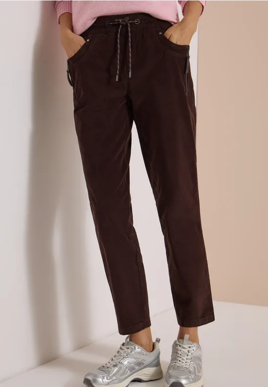 Dunkelbraune Cordhose mit Kordelzug und lässigem geradem Schnitt, kombiniert mit silbernen Sneakers vor beiger Wand.