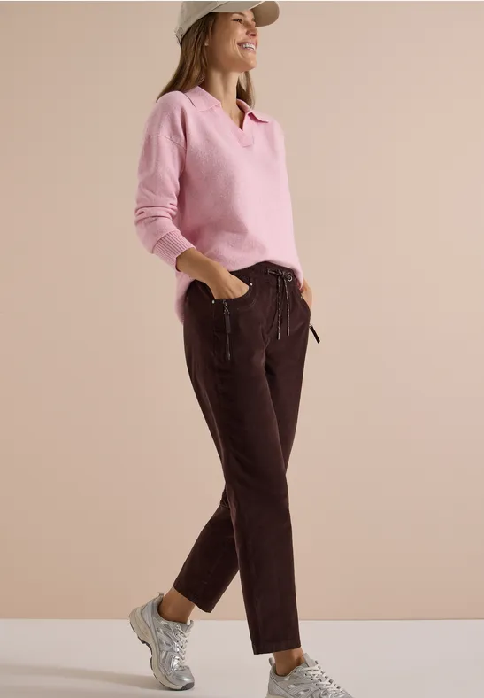 Eine sportliche Frau in rosa Pullover, braunen Hosen und Mütze, die lässig in weißen Sneakern steht.