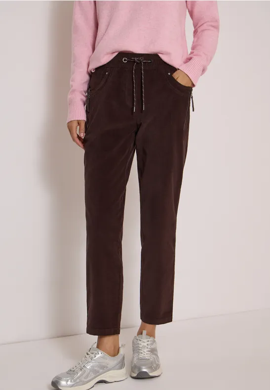 Braune Cordhose mit seitlichen Taschen, locker geschnitten, kombiniert mit rosa Pullover und grauen Sneakern.