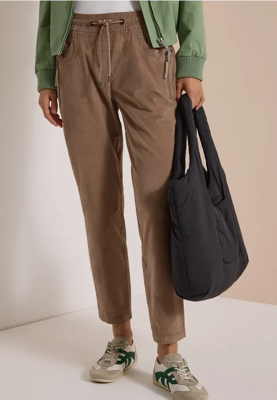 Person trägt braune Cordhose, grüne Jacke, beige Sneaker mit grünen Akzenten und schwarzen Shopper.