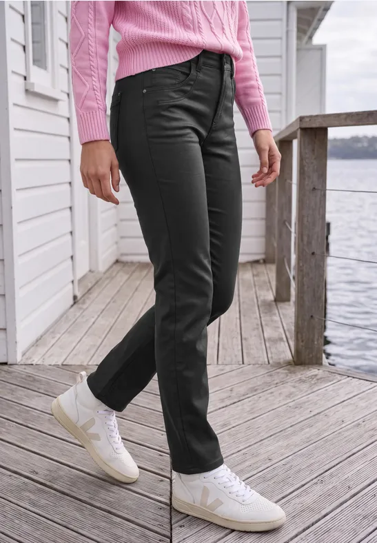 Eine Person steht auf einem hölzernen Steg am Wasser, trägt schwarze Jeans, weiße Sneaker und einen rosafarbenen Pullover.