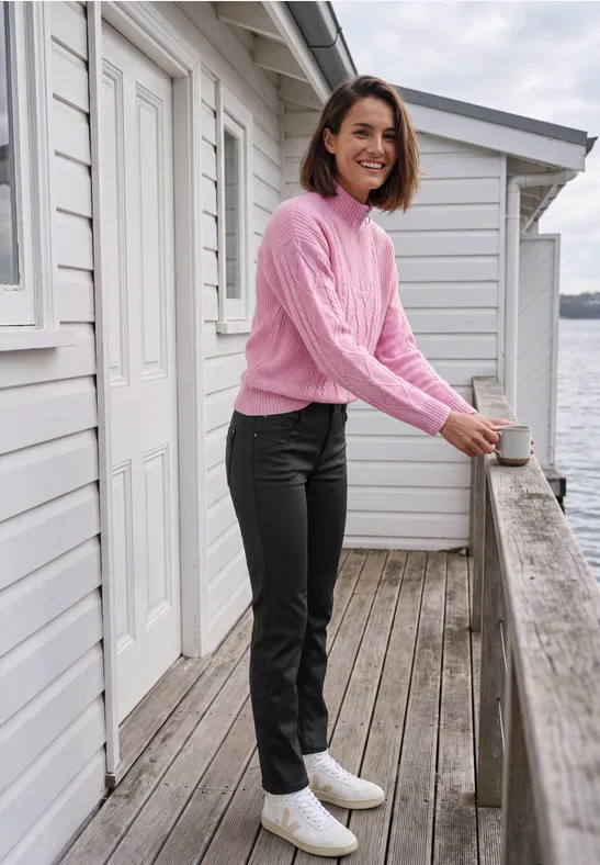 Frau in rosa Pullover, schwarzer Hose und weißen Stiefeletten lehnt auf einer Holzterrasse am Wasser und lächelt.