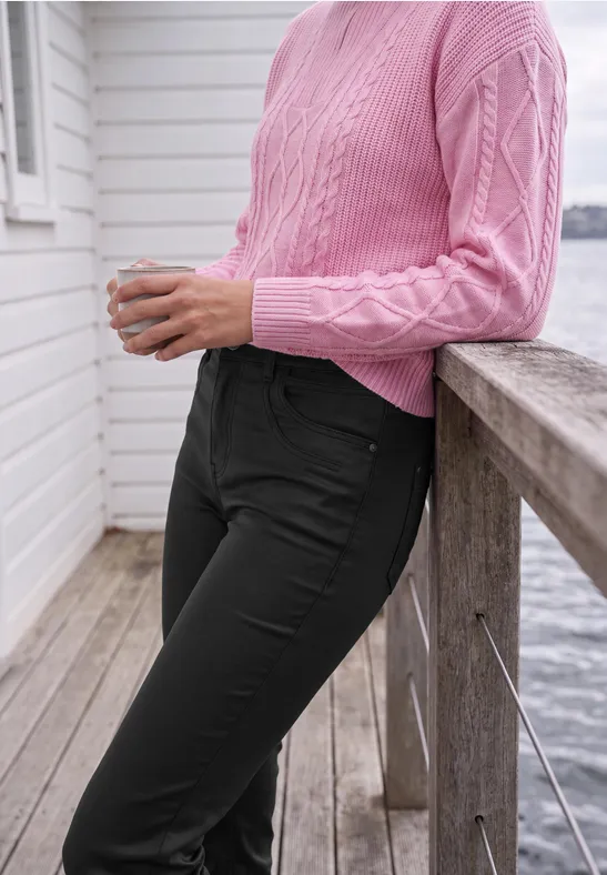 Person lehnt an hölzernem Geländer am Wasser, trägt rosa Strickpulli und schwarze Hose, hält eine Tasse.