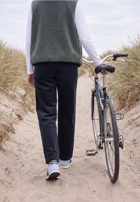Eine Person schiebt auf einem Sandweg zwischen Dünen ein Fahrrad, gesehen von hinten.