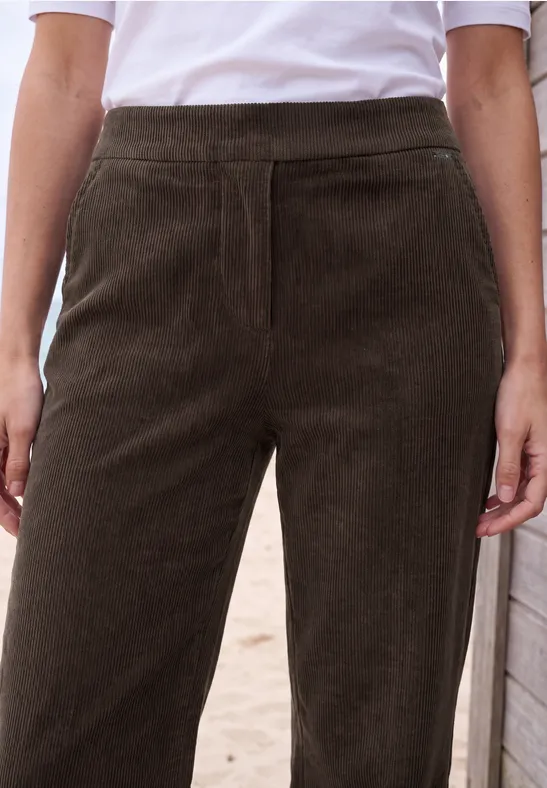 Frau trägt hoch taillierte, dunkelbraune Cordhose mit weißem T-Shirt, Hände locker seitlich.