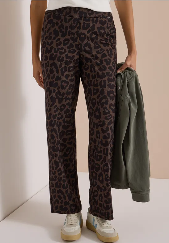 Frau trägt gerade geschnittene Leoprint-Hose und hält eine olivgrüne Jacke.