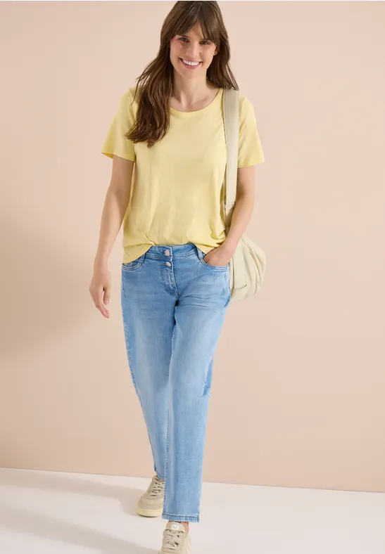 Een vrouw draagt een gele t-shirt, lichte spijkerbroek en sneakers, met een schoudertas tegen neutrale achtergrond.