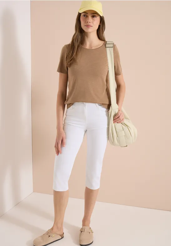 Une femme porte un t-shirt beige, un pantacourt blanc, des chaussures beiges et un sac assorti.