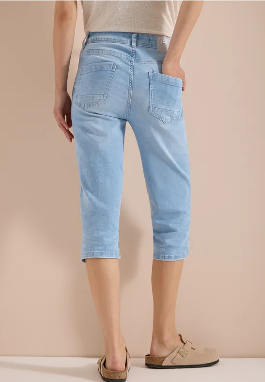 Rückansicht einer Person in hellblauen 3/4-Jeans und beigen Loafers vor neutralem Hintergrund.
