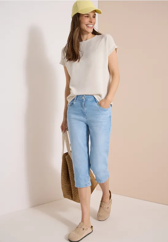 Frau in sommerlicher Kleidung mit hellbeigem T-Shirt, Jeans-Caprihose, Strohtasche und Mokassins
