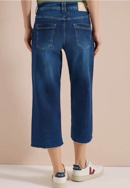 Rückansicht einer Person in dunkelblauen, weiten Cropped-Jeans mit ausgefranstem Saum und weißen Sneakers.