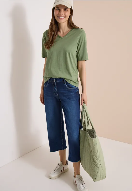 Frau trägt grünes V-Shirt, weite blaue Capri-Jeans, weiße Sneaker und große grüne Tasche vor hellem Hintergrund.