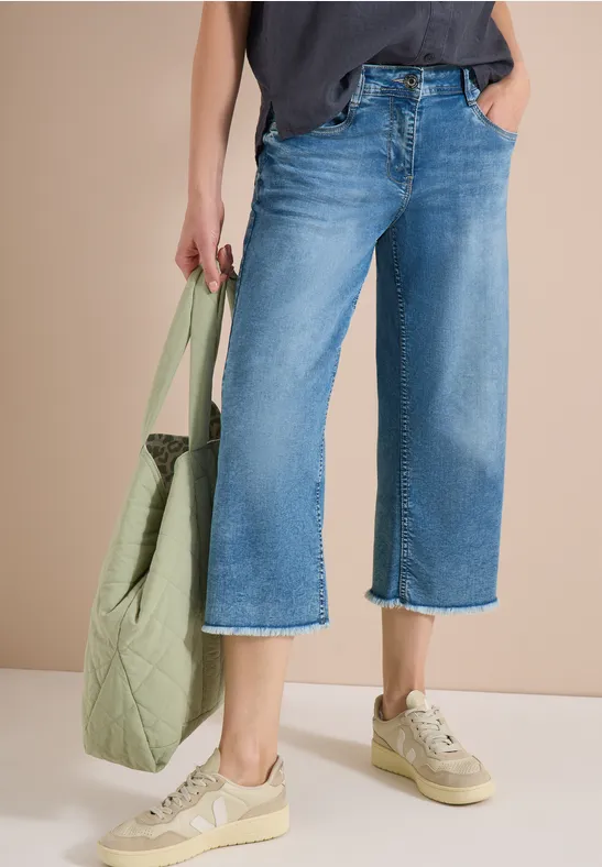 Vrouw in wijde, enkellange jeans met beige sneakers, houdt een groene, doorgestikte tas vast.