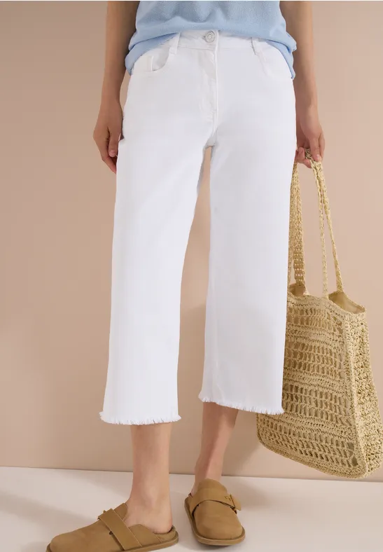 Weiße, weite, knöchellange Jeanshose im Culotte-Stil mit ausgefranster Saum, dazu Basttasche und flache Slipper.