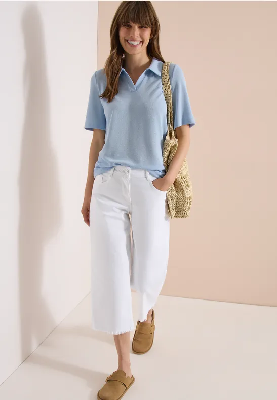 Frau mit hellblauem Poloshirt, weißen Culotte-Hosen, braunen Schuhen und Strohtasche vor schlichter Kulisse.