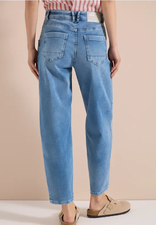 Vrouw van achteren in lichtblauwe high waist-jeans en beige sandalen tegen neutrale achtergrond.