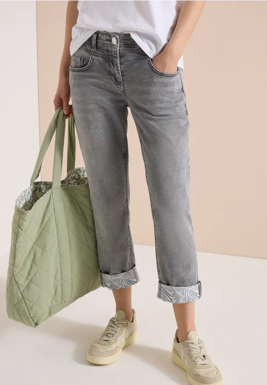 Vrouw draagt grijze omgeslagen spijkerbroek, wit t-shirt, groene tas en beige sneakers.