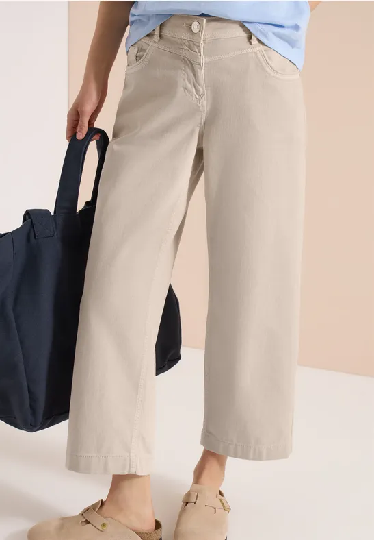 Une personne porte un pantalon large court beige, des baskets claires et tient un sac bleu foncé.