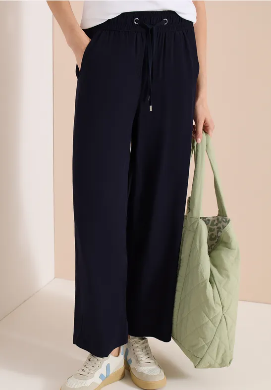 Frau trägt weite schwarze Hose mit Sneakers und hält eine große, gesteppt grüne Tasche.