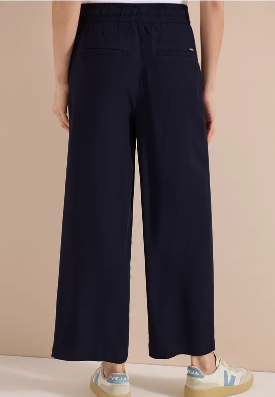 Rückenansicht einer Person in weiten, dunkelblauen Hosen und weißen Sneakern mit grünem Logo.