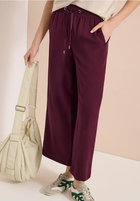 Frau trägt weinrote Jogginghose, weiße Sneaker und hält eine hellbeige Tasche vor neutralem Hintergrund.