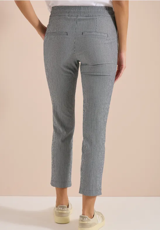 Vrouw van achter in grijze, fijngestreepte 7/8-broek met elastische taille en sneakers, beige achtergrond.