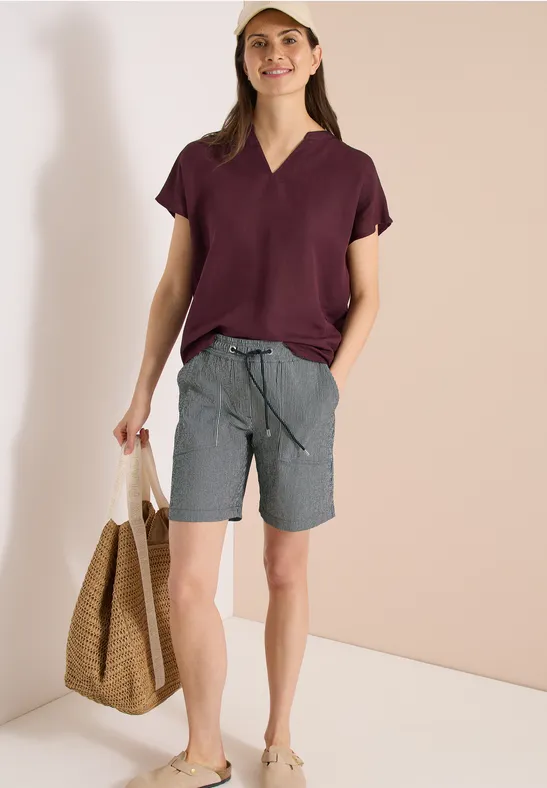 Frau mit weinroter Bluse, grauer Shorts, Strohtasche und Sandalen vor beigem Hintergrund.