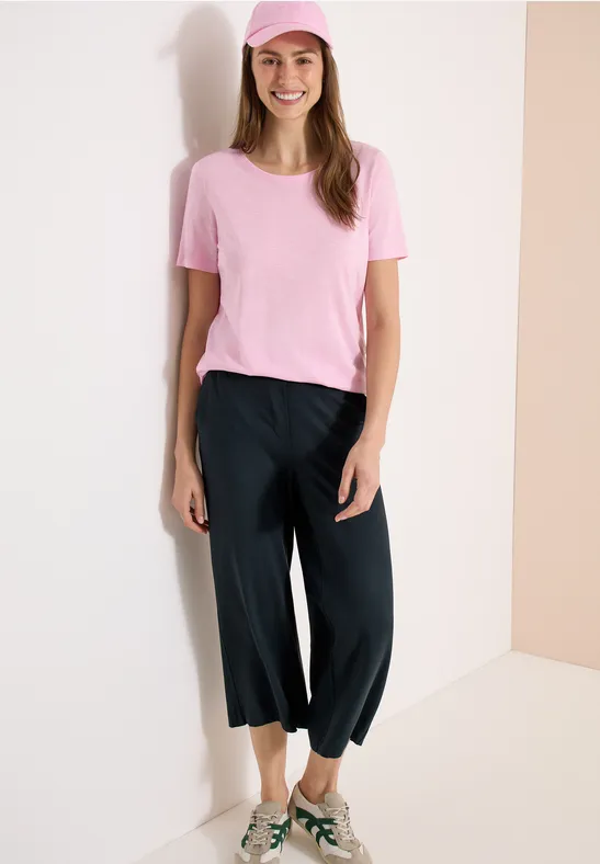 Frau in pinkem T-Shirt und weiten, schwarzen 3/4-Hosen steht vor heller Wand in einer Ecke.