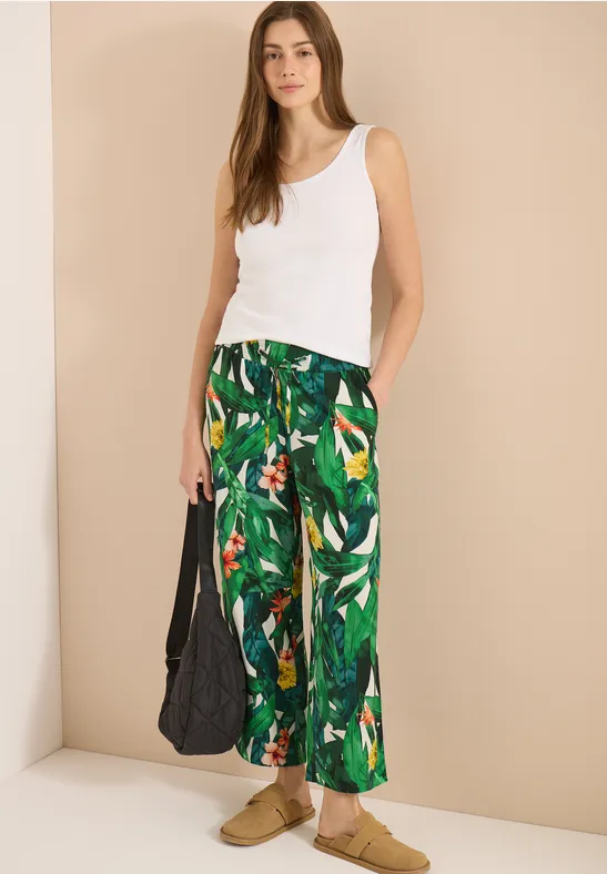 Frau trägt weißes Tanktop, grüne Palmenprint-Hose, beigen Hintergrund, schwarze Tasche und beigen Sandalen.