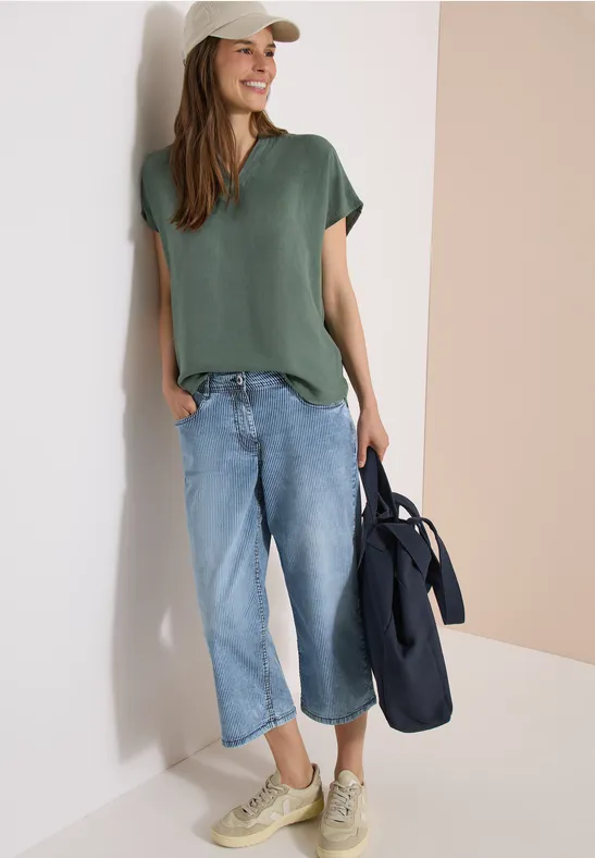 Frau in legerem Outfit: grünes T-Shirt, weite Jeanshose, Sneaker und Tasche lehnt entspannt an Wand.