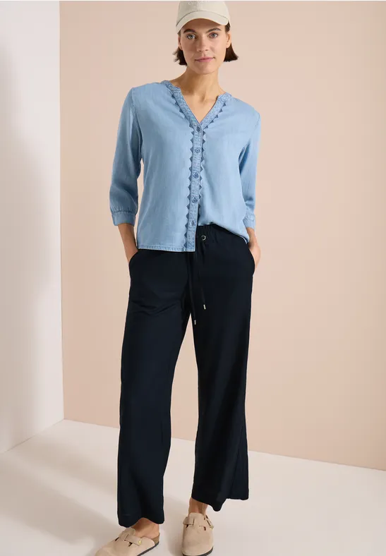Modell trägt hellblaues, vorn geknöpftes Oberteil mit Rüschen und weite schwarze Hosen mit Sneakers.