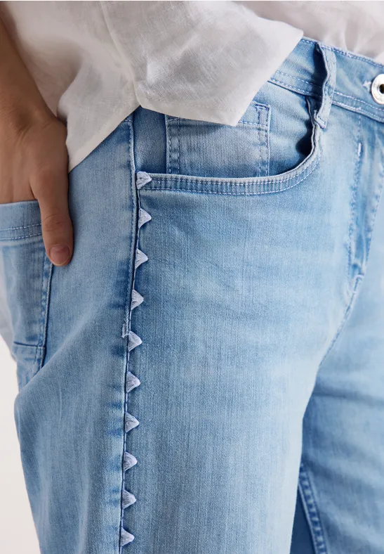 Helle Jeans mit seitlichen Dreieck-Details, weiße Bluse und Hand in Tasche.