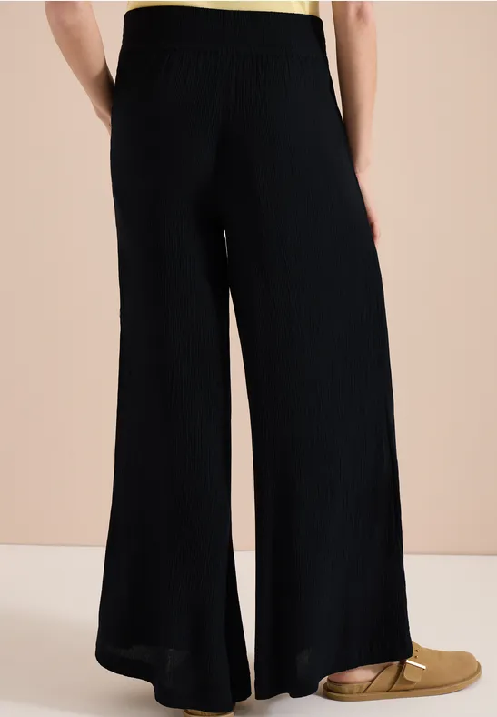 Zwarte, wijde gebreide broek met zijsplit, gecombineerd met gouden, platte instappers tegen een neutrale achtergrond.