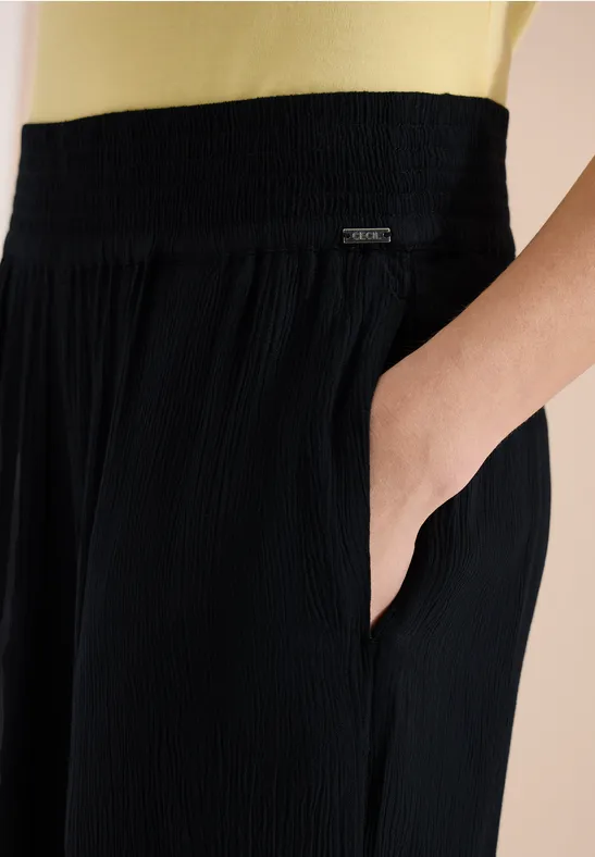 Zwarte, geplooide broek met elastische taille en zijzak, hand in de zak gestoken.