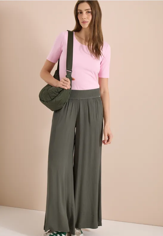 Frau trägt rosa T-Shirt, weite grüne Hose und eine Umhängetasche vor neutralem Hintergrund.