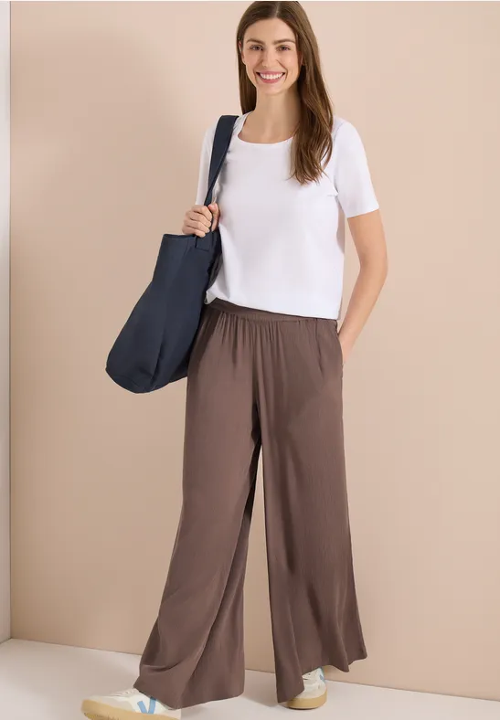 Frau in weißem T-Shirt, brauner weiter Hose, mit blauem Schulterbeutel vor neutralem Hintergrund.