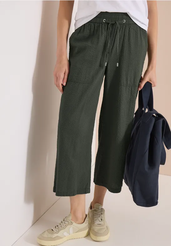 Frau trägt weite, dunkelgrüne 7/8-Hose mit Kordelzug, beige Sneakers und hält eine dunkelblaue Tasche.