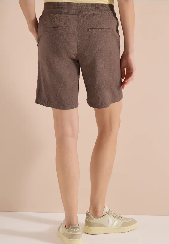 Rückenansicht einer Person in braunen Shorts und beigen Sneakers auf hellem Boden.