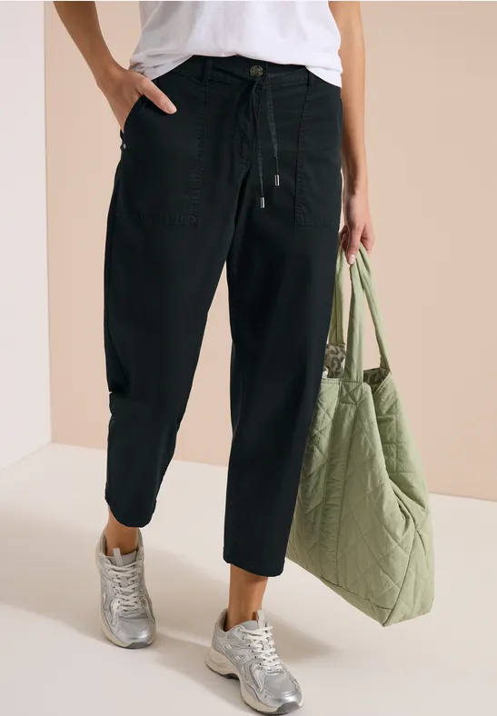 Modell in schwarzer, lässiger Hose, weißem Top, silbernen Sneakern und grünem, gesteppter Shopper.