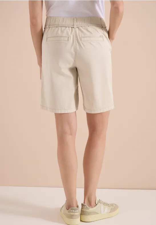 Achteraanzicht van een persoon in lichtbeige korte broek en sneakers tegen beige achtergrond.