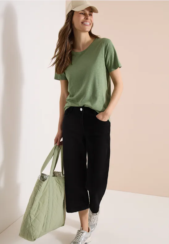 Junge Frau in lockerem grünen T-Shirt, schwarzer Culotte-Hose, weißen Sneakers und großer gesteppten grünen Tasche.