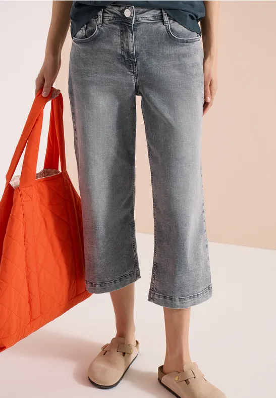 Weite graue 7/8-Jeans, orangene Tasche in der Hand und beige Hausschuhe auf hellem Hintergrund.
