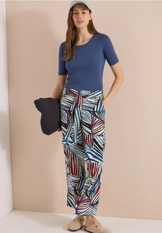Femme en haut bleu, pantalon ample à motifs et espadrilles, tenant un chapeau bleu marine.
