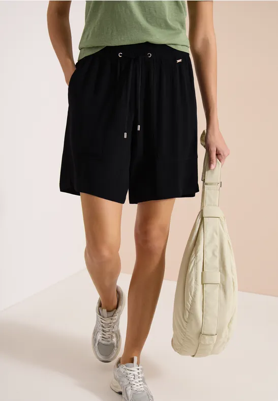Femme en short noir, t-shirt vert, baskets grises et grand sac beige.